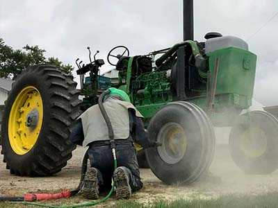 Tractor being sandblasted on site, with blast hose and dust around the wheels